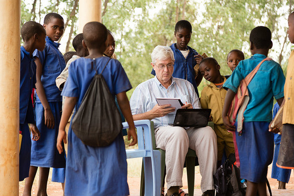 sitting faculty member working on tablet and computer in a group of African children
