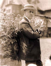An antique portrait of a white man (Rex Beach) wearing a suit and a hat, standing with his back to a bush, his face is facing the camera and body is shifted to the right 