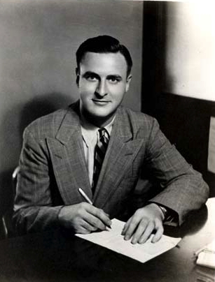 A seated white man, Bucklin Moon, wearing a suit, looking at the camera, while signing a paper document