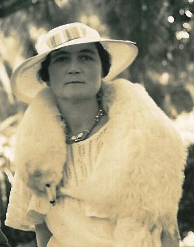 A white woman, Marjorie Kinnan Rawlings, dressed in a white dress, a white fur animal scarf and a white, large brimmed hat standing in front of foliage