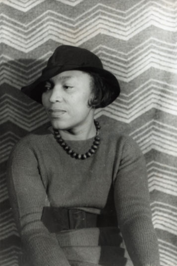 A black woman, Zora Neale Hurston, sitting down against a wall with a chevron-patterned wallpaper, wearing dark clothes and a large brimmed hat, looking down to her right-side