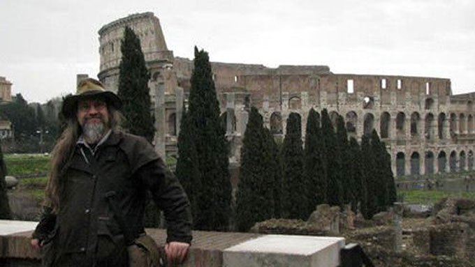 a white, bearded man dressed in coats and a hat, standing in front of the Colosseum