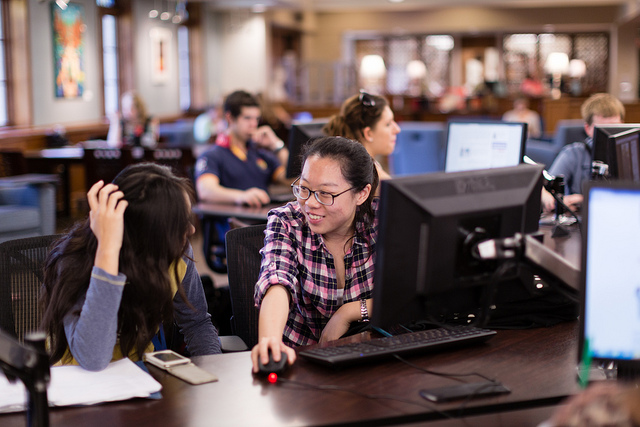 two asian female students looking at each other, seated at a computer